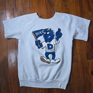 Puede incluir: Sudadera blanca de manga corta con una mascota de dibujos animados azul que sostiene una bandera con la inscripción "DUKE". La mascota lleva una camiseta blanca con una "D" azul. La sudadera tiene cuello redondo y dobladillo acanalado.