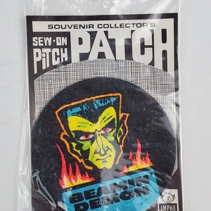 May include: A black and blue sew-on patch with a green demon face with red eyes and flames. The text "Beamin' Demon" is on the patch. The patch is in a plastic bag with the text "Souvenir Collector's Sew-On Patch" on the front.