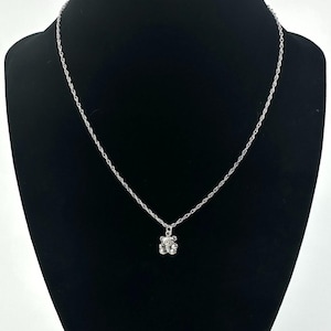 May include: A silver-colored necklace with a small, detailed bear pendant. The pendant is centered on a delicate chain, and the bear has visible features. The necklace is displayed against a black background.