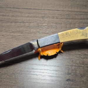 May include: A folding pocket knife with a silver-toned metal blade and a cream-colored handle featuring an etched forest scene. The knife is resting on a small, bright orange plastic crab figurine with black eyes and raised pincers.