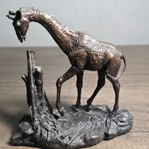 May include: A bronze giraffe sculpture with a textured coat and long neck, standing near a tree stump on a rocky base. The sculpture has a detailed, aged appearance, showcasing the giraffe's distinctive pattern and form. The sculpture is approximately 8 inches tall.