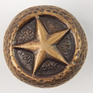 May include: A round bronze-toned metal cabinet knob featuring a raised five-pointed star at the centre. The surface of the knob has a textured, hammered finish, while the recessed areas surrounding the star display a dark, intricate floral pattern.