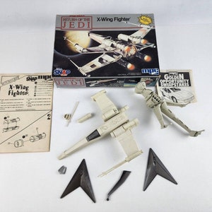 Puede incluir: Un kit de modelo X-Wing Fighter sin montar de la película Star Wars: El Retorno del Jedi. El kit incluye una caja con la imagen del X-Wing Fighter, hojas de instrucciones y varias piezas del modelo en beige y negro.