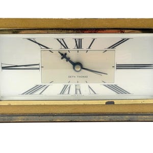 Puede incluir: Reloj rectangular Seth Thomas con marco dorado y esfera blanca. El reloj presenta números romanos y manecillas negras, con el nombre de la marca. Un artículo vintage.