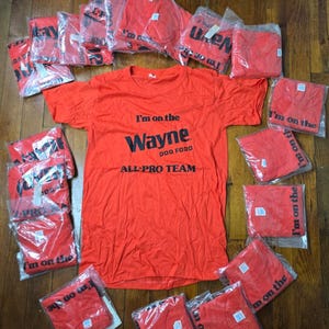 May include: A collection of red t-shirts, some individually packaged, arranged on a wooden surface. The shirts feature the text "I'm on the Wayne Dog Food All-Pro Team" in black. The shirts are a vibrant red color.