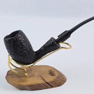 May include: A black briar pipe with a rustic finish, displayed on a wooden stand with a gold metal pipe holder.