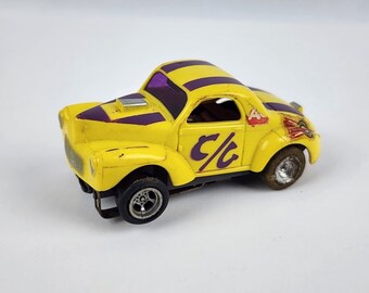 Pode incluir: Um carro de corrida de brinquedo amarelo com listras roxas no teto e no capô. O carro tem as letras "C/c" na lateral e um gráfico de corrida. O carro tem pneus pretos e um detalhe prateado do motor.