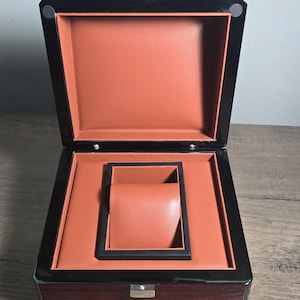 May include: Open wooden watch box with a dark brown exterior and black trim. The interior is lined with a reddish-brown material, and includes a watch holder. The box is open, revealing the interior.