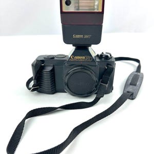 May include: A black Canon T50 film camera with a flash and a black strap. The camera has the Canon logo and model name printed on the front. The flash is a dark red and black colour.