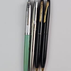 May include: Four vintage pens are displayed against a white background. The pens include a light green pen, a silver pen, and two black pens with gold accents. The pens are of varying lengths and designs, showcasing classic writing instruments.