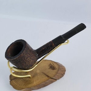 以下が含まれることがあります： 黒いステムが付いた濃い茶色の木製のパイプが、金色の金属製のホルダーが付いた木製のスタンドの上に置かれています