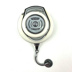 May include: A vintage Beriram Chrostar light meter. The meter is round with a white and silver casing. The face of the meter has the brand name and model name printed on it. A silver chain and disc are attached to the bottom.