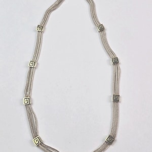 May include: Silver-toned necklace featuring a series of parallel chains. The necklace is adorned with square-shaped decorative elements, each displaying a Greek key design. The necklace is displayed against a plain white background.