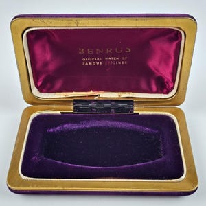 May include: Open vintage watch case with a gold-colored metal frame and a deep purple velvet interior. The case is rectangular with a hinged lid. The text "BENRUS OFFICIAL WATCH OF FAMOUS AIRLINES" is printed inside the lid.