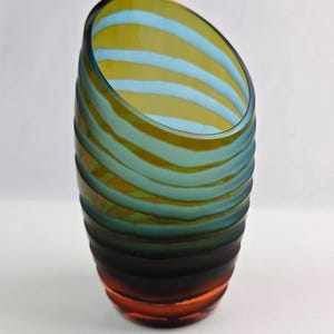 May include: A decorative glass vase with a unique, asymmetrical design. The vase features horizontal stripes in shades of blue, yellow, and orange, creating a gradient effect. The top is angled, adding to its modern aesthetic.