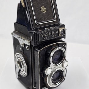 May include: A black Yashica Mat-124 twin-lens reflex camera with a top-mounted viewfinder hood featuring a Y logo. The camera body has textured black leatherette, silver metal accents, two circular lenses, and a winding crank on the side.