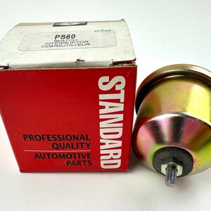 Puede incluir: Un interruptor automotriz nuevo y su caja. La caja es roja con texto blanco que dice "STANDARD" y "PROFESSIONAL QUALITY AUTOMOTIVE PARTS". El interruptor es dorado con una base de goma negra y un perno roscado.