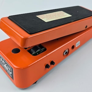 Puede incluir: Pedal de efectos de guitarra naranja con una almohadilla negra para el pie. El pedal tiene un diseño basculante e incluye una entrada y salida jack. La marca "WAH" es visible en el lateral. El pedal está diseñado para guitarras eléctricas.
