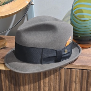 Op de afbeelding: Een grijze vilten fedora hoed met een brede zwarte grosgrain lintband en een decoratieve strik. Aan de zijkant van het lint is een kleine metalen speld bevestigd en achter de strik zit een enkele oranje en bruine veer.
