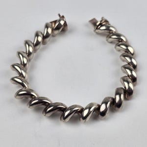 May include: A silver-toned metal bracelet featuring a continuous spiral or twisted rope design. The bracelet is circular in shape with a visible metal clasp mechanism at the top, consisting of a small hook and a rectangular closure component.