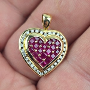 May include: A heart-shaped pendant crafted from yellow gold, featuring a central cluster of square-cut red rubies set with small gold studs. The heart is framed by a border of small, round, clear white diamonds and includes a gold bail at the top.