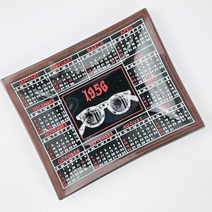 May include: A square, black and red decorative tray featuring a calendar design for the year 1956. The tray has a border with the months of the year and a central image of eyeglasses.