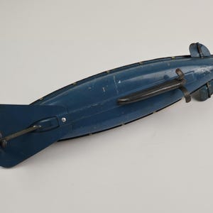 Puede incluir: Un juguete antiguo de dirigible azul con un asa negra y aletas traseras. El juguete tiene un dise&ntilde;o cl&aacute;sico, con un cuerpo largo y aerodin&aacute;mico y una h&eacute;lice en la parte trasera. El dirigible es de metal y muestra signos de desgaste.