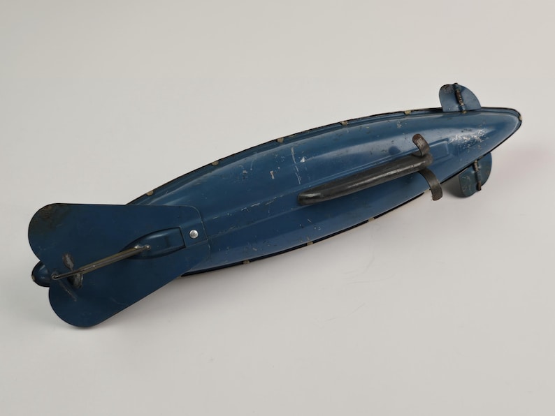 Puede incluir: Un juguete antiguo de dirigible azul con un asa negra y aletas traseras. El juguete tiene un dise&ntilde;o cl&aacute;sico, con un cuerpo largo y aerodin&aacute;mico y una h&eacute;lice en la parte trasera. El dirigible es de metal y muestra signos de desgaste.