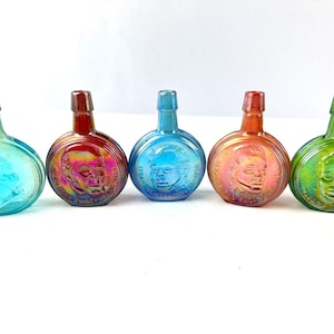 Puede incluir: Cinco pequeñas botellas de vidrio con cuerpos redondos y un solo asa. Cada botella es de un color diferente: azul, rojo, verde, rosa y amarillo. Las botellas tienen una imagen en relieve de un rostro de hombre en la parte delantera.