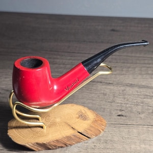 May include: A red Venturi tobacco pipe with a black stem, resting on a gold-colored stand. The pipe is displayed on a small, round wooden base. The word "Venturi" is printed on the side of the pipe.