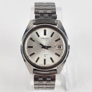May include: A silver Seiko automatic wristwatch with a silver band. The watch face has black hour and minute hands, a black second hand, and a silver date window at the 3 o'clock position. The watch face also has the text "SEIKO AUTOMATIC" and "17 JEWELS".