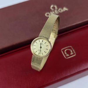 May include: A gold-tone Omega De Ville quartz wristwatch featuring a round cream-colored dial with thin black hour markers and a matching gold-tone mesh metal bracelet. The watch face displays the Omega logo and the text De Ville Quartz.