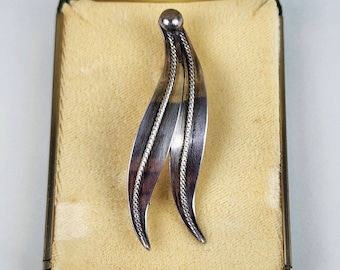 May include: A silver brooch in the shape of two stylized leaves, with a textured design. The brooch is displayed in a beige velvet box with a green lining.