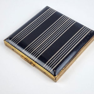 May include: A black and white striped compact with a gold metal frame. The compact has a hinged lid and a small latch.