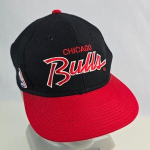 Puede incluir: Gorra de béisbol negra y roja con el logotipo de los Chicago Bulls bordado en rojo en la parte delantera. La gorra tiene una visera roja y una corona negra.