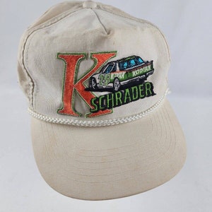 May include: A white baseball cap with a rope trim. The front of the cap features an embroidered patch with a large orange "K" and a green race car with the text "KODIAK SCHRADER".
