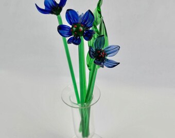 May include: A glass floral arrangement featuring three blue flowers with green stems and leaves in a clear glass vase. The flowers have a detailed design with red accents in the center. The vase has a classic, elegant shape.