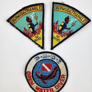 May include: Two triangular embroidered patches featuring a red devil diver with a trident, yellow flames, and the text BERMUDA TRIANGLE 50 DIVES, alongside one circular PADI Open Water Diver patch with a blue globe, red diver, and white flag.
