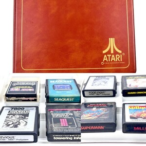May include: A selection of Atari 7800 video game cartridges, including "Seaquest", "Food Fight", and "Superman", presented with a brown Atari case. The Atari logo is visible on the case. The cartridges are in a white tray.