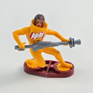 May include: A yellow plastic action figure of a person wearing a yellow spacesuit with a red and white design on the chest. The figure is holding a silver weapon and is standing on a red base.