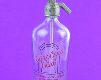 May include: A clear glass bottle with a metal soda siphon top. The bottle has a red and black label that reads "Carolina Club" and "Carolina Coca Cola Bottling Co Du Bois PA". The label also says "Bottled to be Chilled before using".