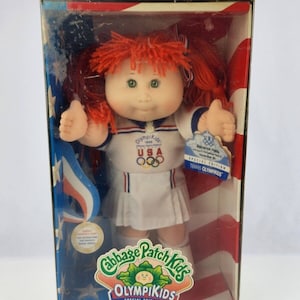 May include: Cabbage Patch Kids Olympikids Special Edition doll in a white and blue athletic outfit with the USA flag and Olympic rings. The doll has red hair and green eyes. The box is red, white, and blue with the Cabbage Patch Kids logo.