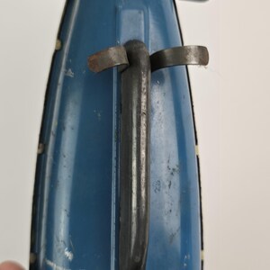 Puede incluir: Primer plano de un barco de juguete azul vintage con un asa negra y detalles met&aacute;licos. El barco muestra signos de desgaste, con ara&ntilde;azos y pintura descolorida, lo que sugiere que es un art&iacute;culo antiguo o muy querido. El dise&ntilde;o incluye peque&ntilde;as aletas curvadas hacia arriba.