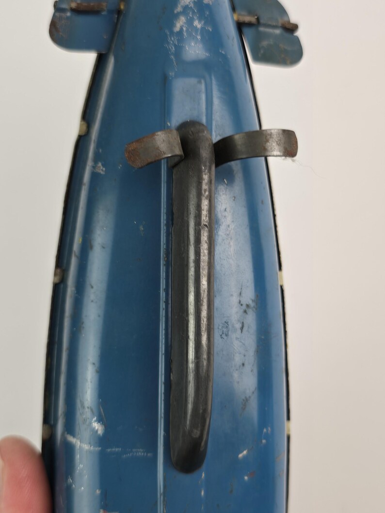 Puede incluir: Primer plano de un barco de juguete azul vintage con un asa negra y detalles met&aacute;licos. El barco muestra signos de desgaste, con ara&ntilde;azos y pintura descolorida, lo que sugiere que es un art&iacute;culo antiguo o muy querido. El dise&ntilde;o incluye peque&ntilde;as aletas curvadas hacia arriba.