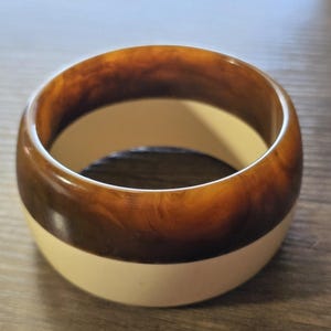 May include: A wide, round bangle bracelet with a two-tone design. The top portion is a marbled brown, while the bottom is a solid cream color. The bracelet is made of a smooth, glossy material.