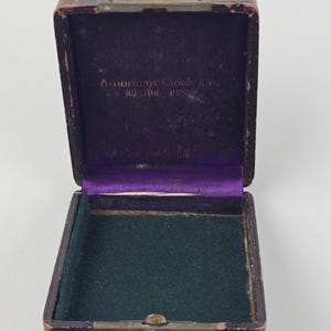 May include: An open, antique, rectangular box with a hinged lid. The exterior is dark brown with a worn texture. The interior features a purple velvet lining on the lid and a green felt base. The text "American Clock Co. Boston, Mass." is visible inside the lid.