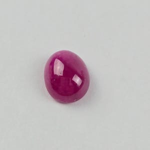 May include: A polished, oval-shaped ruby gemstone. The gem is a deep, saturated pinkish-red color with a smooth, reflective surface. The stone is set against a plain white background.