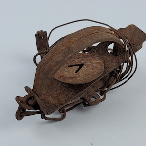 May include: A rusty metal animal trap with a number "7" stamped on it. The trap is closed and has a chain attached to it.