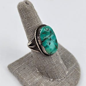 May include: A silver ring featuring an oval-shaped turquoise stone with dark matrix veining. The stone is set in a smooth bezel surrounded by a twisted rope border. The band is constructed from three thin silver wires that merge into one shank.