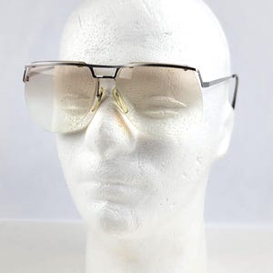 May include: Vintage sunglasses with a silver frame and gradient lenses. The sunglasses have a rectangular shape and a bridge over the nose. The lenses transition from a light color at the top to a darker shade at the bottom.